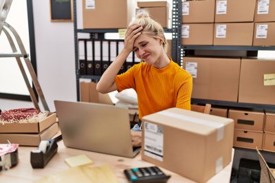 Young blonde woman working at small business ecommerce using laptop stressed with one hand on head, surprised and angry face