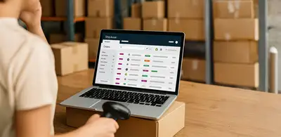 Warehouse worker using a laptop with ShipAccel ecommerce software to track and process online orders, surrounded by cardboard boxes.